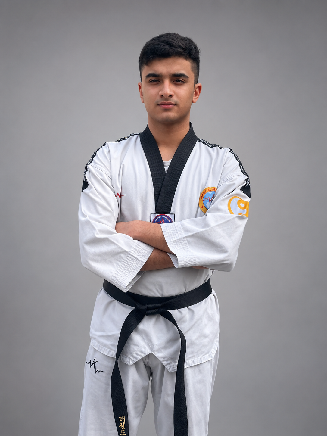Arjun Basandrai standing with arms crossed in a white taekwondo dobok and black belt against a plain grey studio background